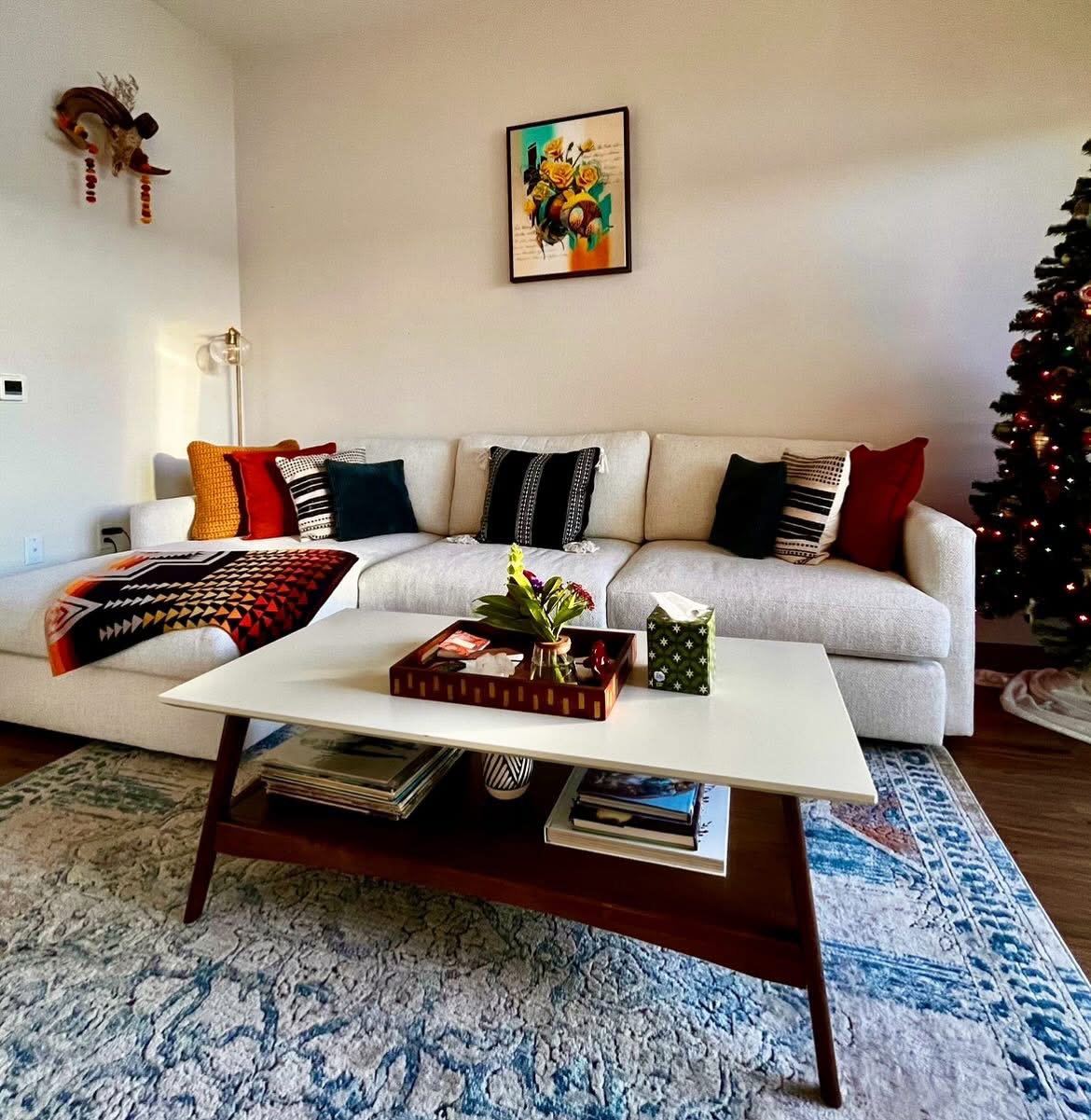 A modern living room with a white sectional sofa, assorted throw pillows, a coffee table with decor, and a decorated Christmas tree in the corner.